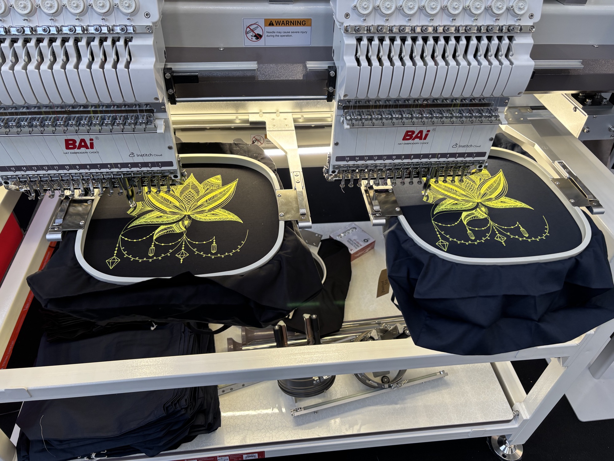 Two heads of a multi-head embroidery machine stitching the same yellow lotus design onto navy garments, with a stack of the same garments waiting below.