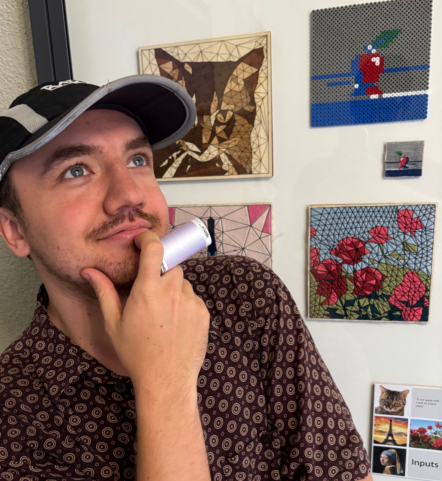 A maker in a cap holding a spool of thread to his chin, looking up in thought, in front of a wall of finished embroidered pieces.