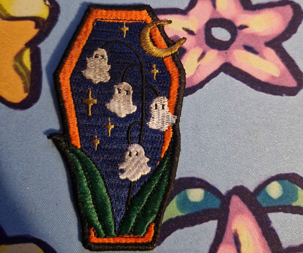 A coffin-shaped patch embroidered with white ghosts, a gold crescent moon and green leaves on navy.
